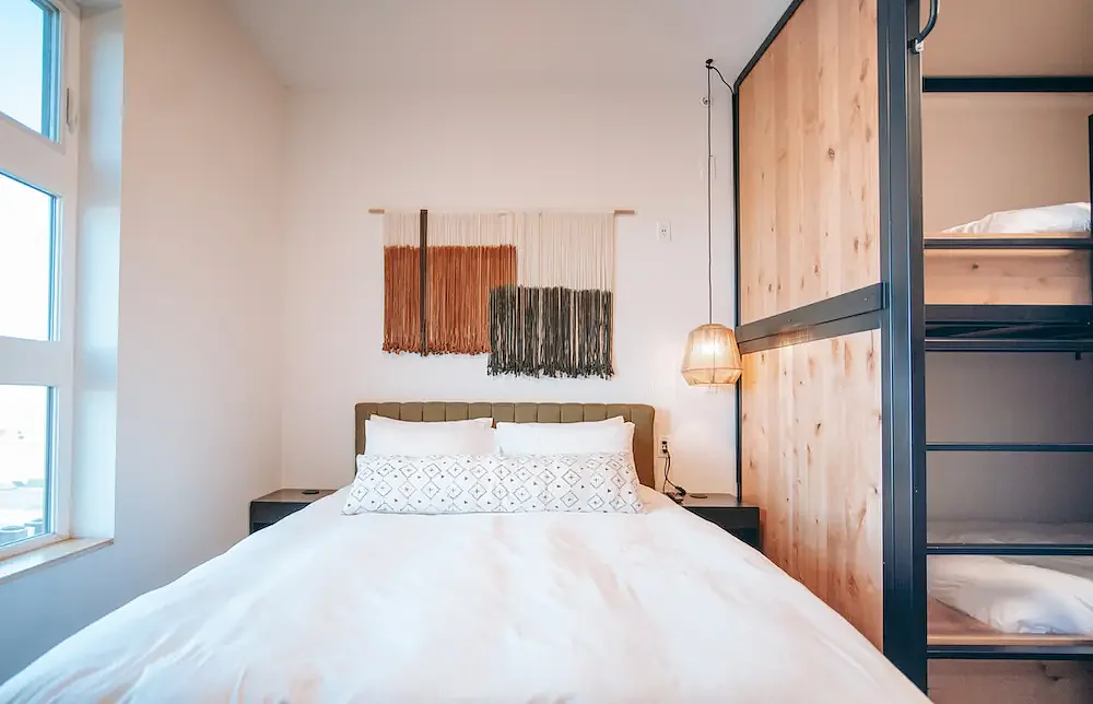A neatly made bed with white linens and pillows, with bunk beds to the right and a wall hanging above the bed.